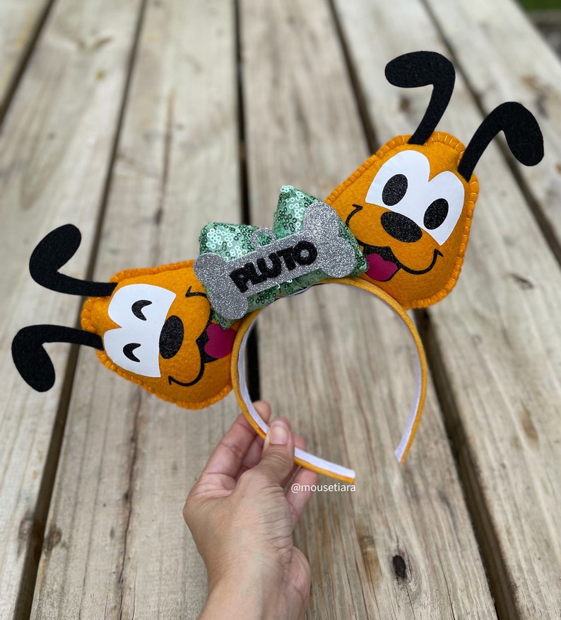 Peut inclure: Un serre-t&ecirc;te oreilles de Pluto en feutre orange avec un n&oelig;ud vert et argent&eacute; qui dit "Pluto" dessus.