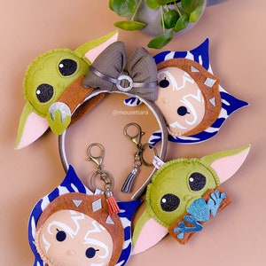 May include: Three felt character keychains featuring Baby Yoda, Ahsoka Tano, and a brown and blue character. The keychains are attached to silver key rings with tassels. The keychains are on a light brown surface.