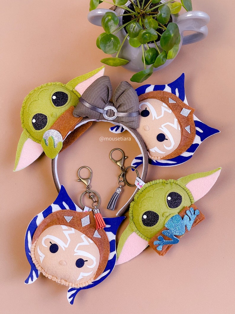 May include: Three felt character keychains featuring Baby Yoda, Ahsoka Tano, and a brown and blue character. The keychains are attached to silver key rings with tassels. The keychains are on a light brown surface.