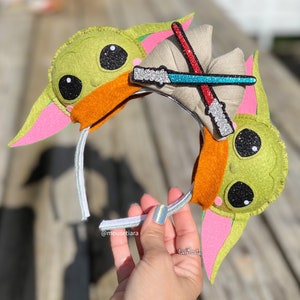 May include: Green and pink felt character ears with a sparkly blue and red lightsaber headband. The headband is attached to a white headband. The ears are shaped like a character from the Star Wars franchise. The text "@mousetiara" is visible on the headband.
