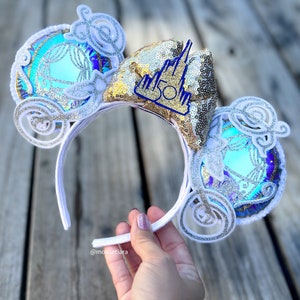 May include: White headband with a gold and blue Cinderella carriage design. The carriage is decorated with glitter and sequins. The headband also features a gold castle charm with the number 50 on it.
