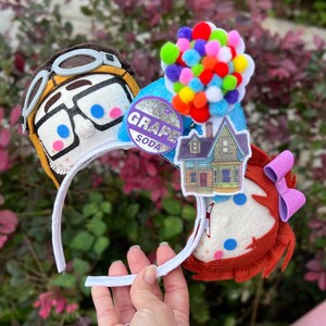 May include: A pair of white mouse ears with a felt character of a young boy with glasses and a felt character of a young girl with a pink bow. The boy character has a grape soda logo on his ear. The girl character has a house with balloons on her ear.