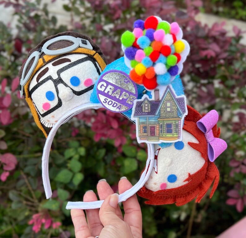 May include: A pair of white mouse ears with a felt character of a young boy with glasses and a felt character of a young girl with a pink bow. The boy character has a grape soda logo on his ear. The girl character has a house with balloons on her ear.