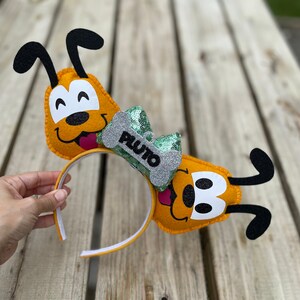 Peut inclure: Une paire d'oreilles de Pluto en feutre jaune avec des oreilles en feutre noir et un n&oelig;ud en sequins vert et argent avec une &eacute;tiquette en forme d'os qui dit "Pluto".