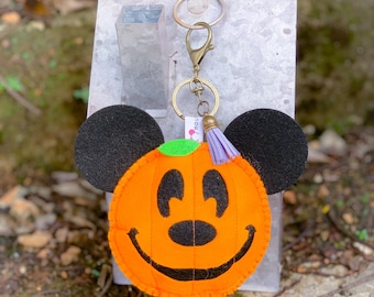 Pode incluir: Chaveiro de abóbora laranja de feltro com orelhas pretas do Mickey Mouse e um rosto sorridente de Jack-o'-lantern. O chaveiro tem um fecho e um anel dourados e uma borla roxa. O texto "@mousetiara 2020" está na parte inferior.