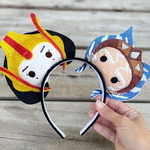 May include: A pair of character-themed headband ears with a black headband. The ears are made of felt and feature a yellow and red character on the left and a blue and brown character on the right.