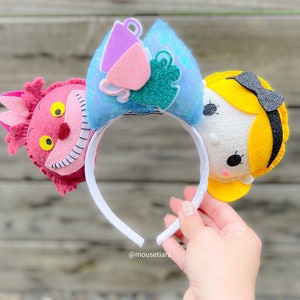 Può includere: Un paio di orecchie di topo in feltro con un design a tazza da tè blu e verde su un orecchio e un design di Gatto del Cheshire rosa e rosso sull'altro orecchio. Le orecchie sono attaccate a un cerchietto bianco.