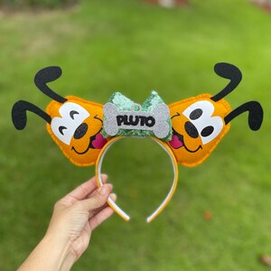 Peut inclure: Un serre-t&ecirc;te oreilles de chien Pluto en feutre jaune avec un n&oelig;ud en paillettes vert et argent et une &eacute;tiquette en forme d'os qui dit "PLUTO".