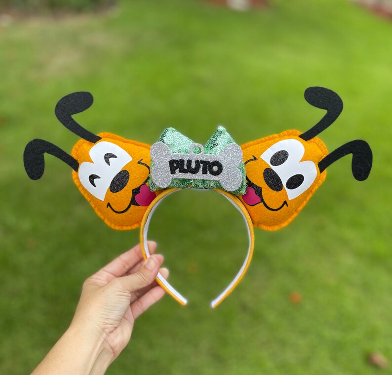 Peut inclure: Un serre-t&ecirc;te oreilles de chien Pluto en feutre jaune avec un n&oelig;ud en paillettes vert et argent et une &eacute;tiquette en forme d'os qui dit "PLUTO".