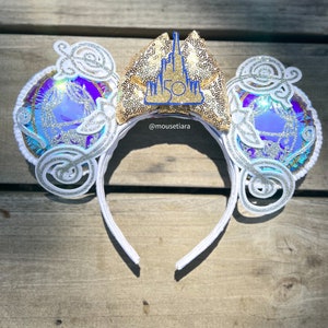 May include: White Minnie Mouse ears with a gold sequin bow featuring a blue castle with the number 50 on it. The ears have a white and iridescent design with swirls. The text '@mousetiara' is visible on the bow.
