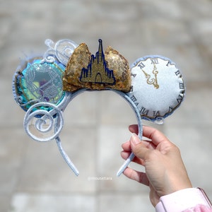 May include: A light blue and silver headband with decorative ears. One ear features a clock design, and the other has a carriage design. The centre of the headband has a gold sequined bow with a blue castle emblem.