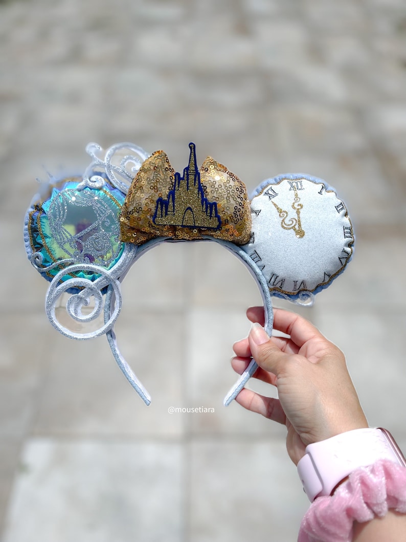 May include: A light blue and silver headband with decorative ears. One ear features a clock design, and the other has a carriage design. The centre of the headband has a gold sequined bow with a blue castle emblem.