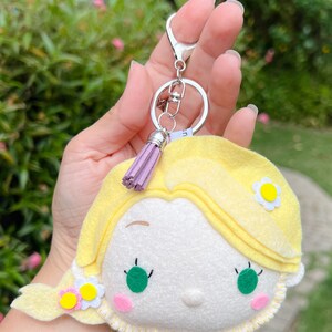 Peut inclure: Un porte-clés en feutre jaune représentant un personnage de dessin animé avec des cheveux blonds, des yeux verts et un fard à joues rose. Le porte-clés a un fermoir argenté et un pompon violet. Le personnage a une fleur sur le côté de la tête.