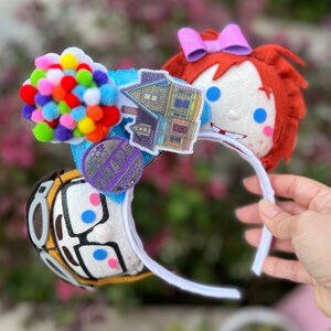 May include: A pair of white mouse ears with a colorful balloon house and a purple Grape Soda pin on one ear and a red-haired girl with blue eyes on the other ear.