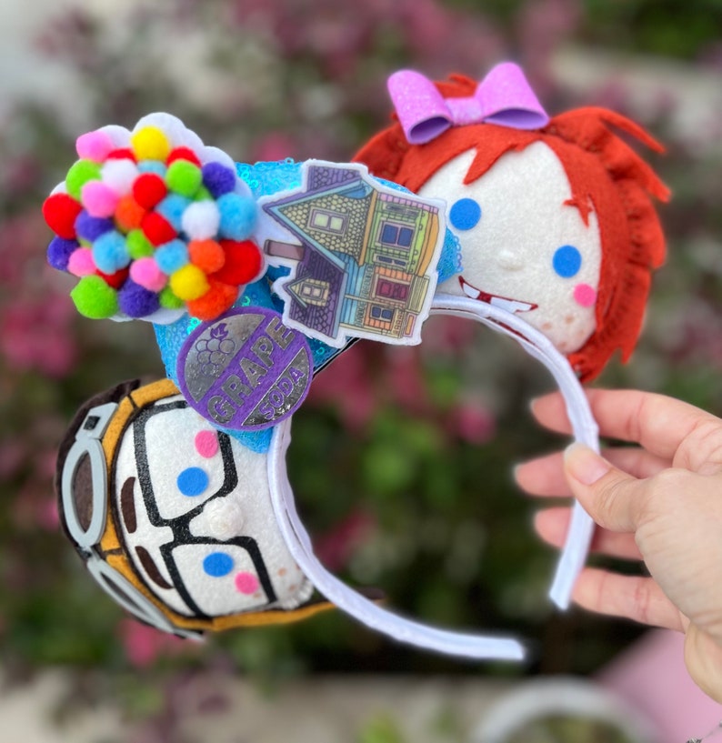 May include: A pair of white mouse ears with a colorful balloon house and a purple Grape Soda pin on one ear and a red-haired girl with blue eyes on the other ear.