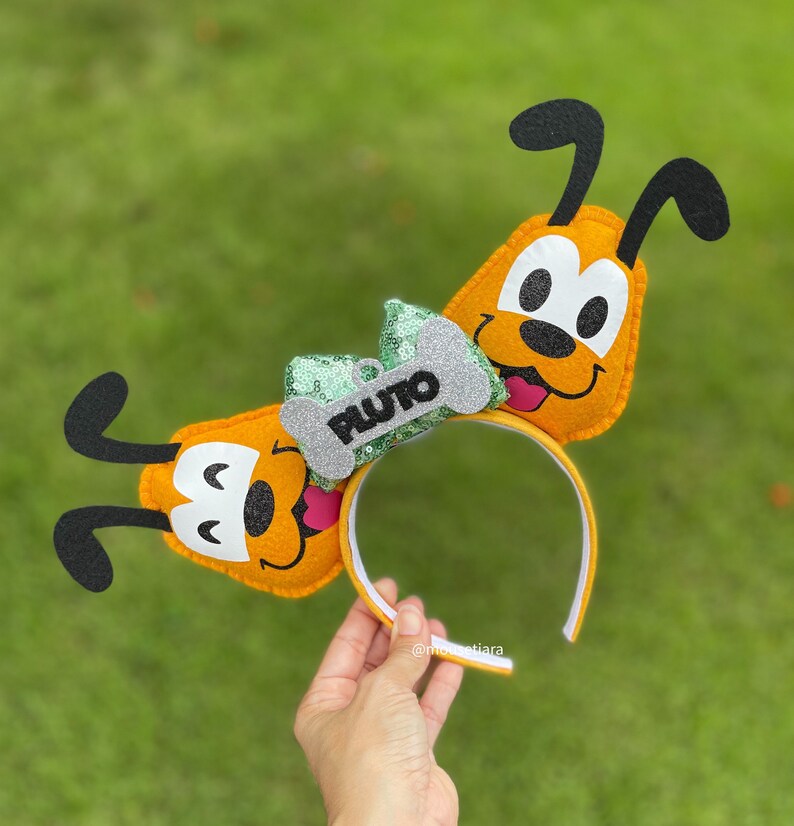 Peut inclure: Un serre-t&ecirc;te en feutre jaune avec deux oreilles de Pluto. Les oreilles sont en forme de t&ecirc;te de Pluto et ont un nez noir et des yeux blancs. Le serre-t&ecirc;te a un n&oelig;ud vert et argent avec une &eacute;tiquette en forme d'os qui dit "PLUTO".