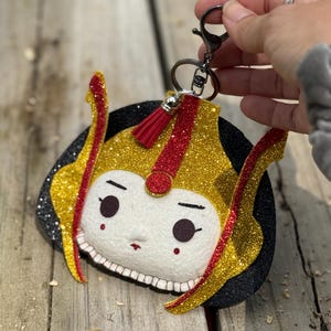 May include: A gold and red glitter felt keychain with a cartoon face and a black felt base. The keychain has a silver metal ring and a black metal clasp.