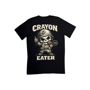 Könnte beinhalten: Schwarzes T-Shirt mit einem Cartoon-Schädel in Militäruniform und Helm, der Buntstifte hält. Der Text "CRAYON EATER" befindet sich über und unter der Grafik. Ein US-Emblem befindet sich auf der rechten Seite des Schädels.