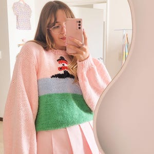 Puede incluir: Un suéter rosa claro con un diseño de faro, con mangas rosas, una sección azul y verde y una falda plisada rosa. El suéter tiene cuello redondo y un corte holgado.