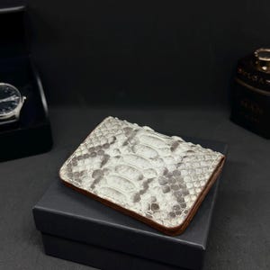 Pode incluir: Uma carteira retangular com um padrão de pele de cobra em tons de branco e cinza, com bordas marrons. A carteira está em cima de uma caixa preta, com um relógio e um frasco de colônia no fundo.