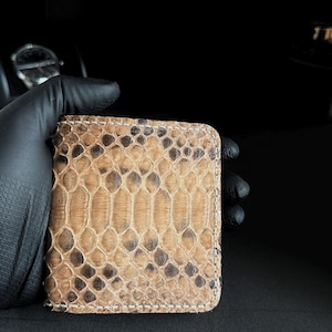 May include: A rectangular wallet with a snakeskin pattern in shades of tan, brown, and black. The wallet is held by a gloved hand against a dark background. The edges are stitched with a light-colored thread.