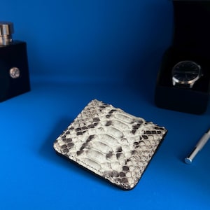 May include: A square, bi-fold wallet with a snakeskin pattern in shades of gray and white. The wallet is on a blue surface, with a black perfume bottle and a watch in a black box in the background.