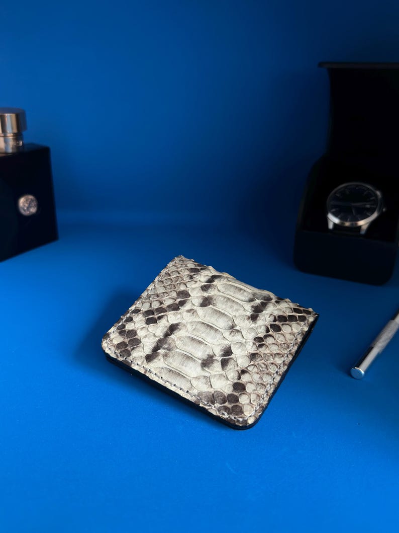 May include: A square, bi-fold wallet with a snakeskin pattern in shades of grey and white. The wallet is on a blue surface, with a black perfume bottle and a watch in a black box in the background.