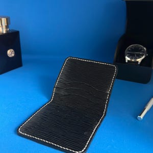 May include: Black leather wallet with white stitching, open to show card slots. A black watch in a box, a bottle of cologne, and a silver pen are also visible against a blue background. The wallet has a textured surface.