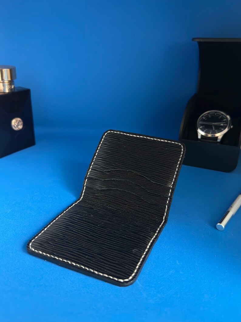 May include: Black leather wallet with white stitching, open to show card slots. A black watch in a box, a bottle of cologne, and a silver pen are also visible against a blue background. The wallet has a textured surface.