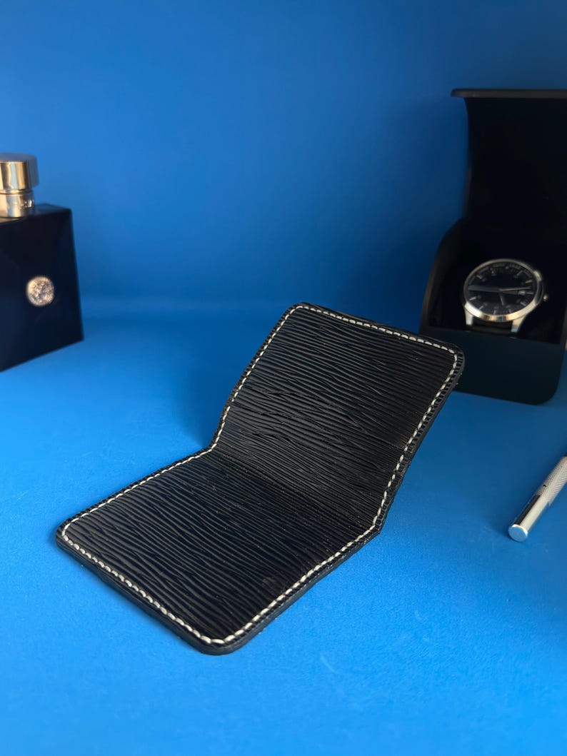 May include: Black leather wallet with white stitching, open to show the inside. The wallet is on a blue surface, with a watch in a black box and a bottle of cologne in the background.