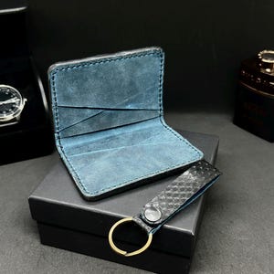 May include: A handmade leather wallet with a blue interior and a matching keychain. The wallet is open, revealing card slots. The keychain has a textured pattern and a gold-colored ring. The items are displayed on a black box.