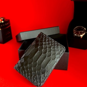 May include: A black, textured leather wallet with a reptile-like pattern, inside an open black box. The wallet is the focal point against a vibrant red background. A bottle of cologne and a watch are visible in the background.