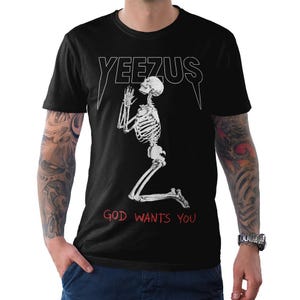 May include: Black t-shirt featuring the word "YEEZUS" in white, above a kneeling skeleton illustration. The text "GOD WANTS YOU" is printed in red below the skeleton. The shirt has short sleeves.
