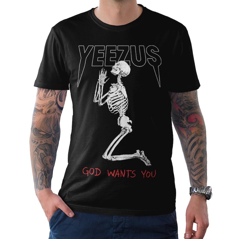 May include: Black t-shirt featuring the word "YEEZUS" in white, above a kneeling skeleton illustration. The text "GOD WANTS YOU" is printed in red below the skeleton. The shirt has short sleeves.