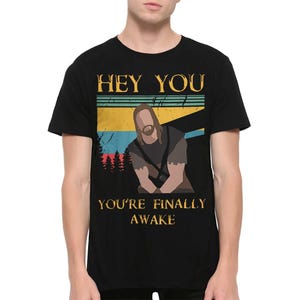 Puede incluir: Camiseta negra con el texto "HEY YOU" sobre un gráfico de una persona con pelo largo y barba. Debajo del gráfico, el texto dice "YOU'RE FINALLY AWAKE". El diseño incluye un esquema de color retro con rayas azules, amarillas y rojas.