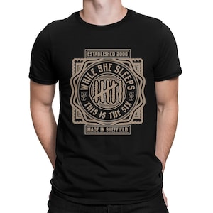 May include: Black t-shirt with a circular graphic in tan and white. The graphic includes the text "WHILE SHE SLEEPS", "THIS IS THE SIX", "ESTABLISHED 2006", and "MADE IN SHEFFIELD". The shirt is short-sleeved and has a crew neck.