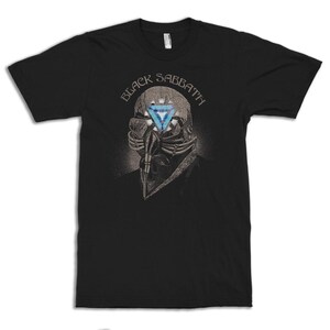May include: Black t-shirt featuring a detailed illustration of a skeletal figure with a blue triangle in place of an eye. The text "BLACK SABBATH" is arched above the figure in a stylized font. The design is centered on the shirt.
