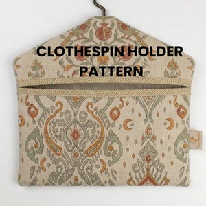 Puede incluir: Un patrón de soporte para pinzas de ropa beige con un estampado decorativo de flores y pájaros estilizados. El soporte tiene una abertura superior y está suspendido de una percha de metal. El texto "CLOTHESPIN HOLDER PATTERN" está impreso en la parte delantera.