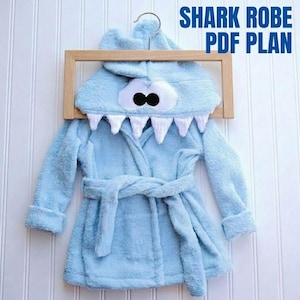 May include: Light blue shark-themed bathrobe with a hood featuring large white teeth and black eyes. The robe has a matching belt and is hanging on a wooden hanger. Text on the image reads "SHARK ROBE PDF PLAN".