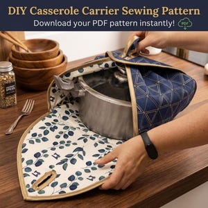 May include: A fabric casserole carrier with a navy blue exterior featuring a gold geometric triangle pattern and a cream-coloured interior lining with a blue eucalyptus leaf print. The carrier has gold trim and a handle, shown holding a metal pot.