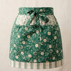 May include: A green floral apron with a white and beige striped bottom hem. The apron features a white floral pocket and a matching tie. The main fabric has a pattern of white flowers and leaves.
