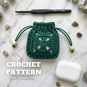 May include: A dark green crocheted drawstring pouch with a floral design, wooden beads, and a black crochet hook. The text "CROCHET PATTERN PDF DOWNLOAD" is displayed below the pouch, along with a white case.