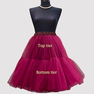 May include: A two-tiered, magenta-colored tulle petticoat displayed on a mannequin. The top tier is labeled, and the bottom tier is fuller. The waistband is a dark brown color. A pearl necklace is around the mannequin's neck.