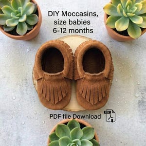Puede incluir: Un par de mocasines de ante marrón para bebé con flecos, talla 6-12 meses. El texto dice: DIY Moccasins, size babies 6-12 months, PDF file Download. Los mocasines están sobre una base de madera rodeados por tres plantas suculentas en macetas.