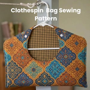 May include: A patterned clothespin bag sewing pattern. The bag features a colorful design with orange, blue, and teal geometric shapes. The interior lining is a brown and tan checkered pattern. The bag is hanging on a white hanger.