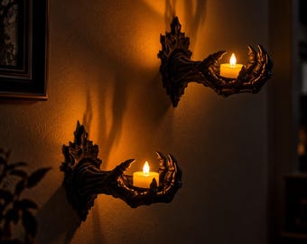 May include: Two dark, claw-like wall sconces mounted on a wall, each featuring a sculpted hand extending outwards to hold a small, glowing yellow tea light candle. The sconces have a textured, ornate base and cast long, dramatic shadows on the wall.