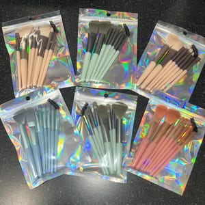 May include: Several sets of makeup brushes, each in a clear, iridescent plastic bag. The sets feature brushes with handles in various colours, such as beige, pale blue, and pink. These brushes are designed for applying makeup.