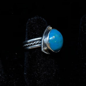 Puede incluir: Un anillo de plata con una piedra grande, redonda y azul brillante. El anillo tiene una banda trenzada y un engaste decorativo con pequeños detalles dorados. El anillo se muestra sobre un fondo negro.