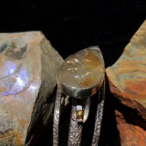 May include: A silver ring with a teardrop-shaped, clear gemstone containing golden inclusions. The ring's band features intricate floral and swirl patterns. A small gold accent is visible near the gemstone.