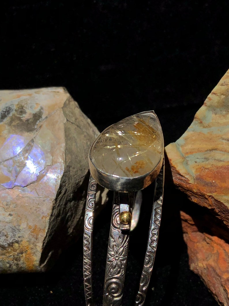 May include: A silver ring with a teardrop-shaped, clear gemstone containing golden inclusions. The ring's band features intricate floral and swirl patterns. A small gold accent is visible near the gemstone.